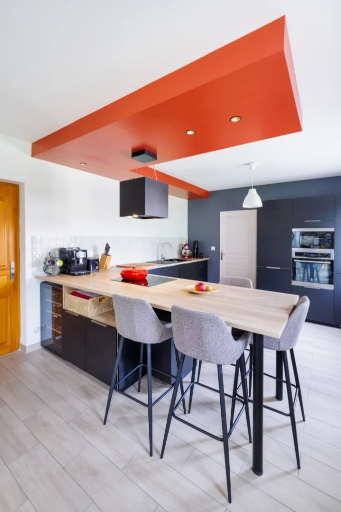 Cuisine moderne ouverte avec un grand îlot central en bois clair équipé d'une plaque à induction et d'une hotte suspendue noire. L'espace repas comprend trois chaises hautes grises. Le design se distingue par un coffrage de plafond rouge vif avec spots intégrés, des meubles gris anthracite et un mur d'accent bleu foncé