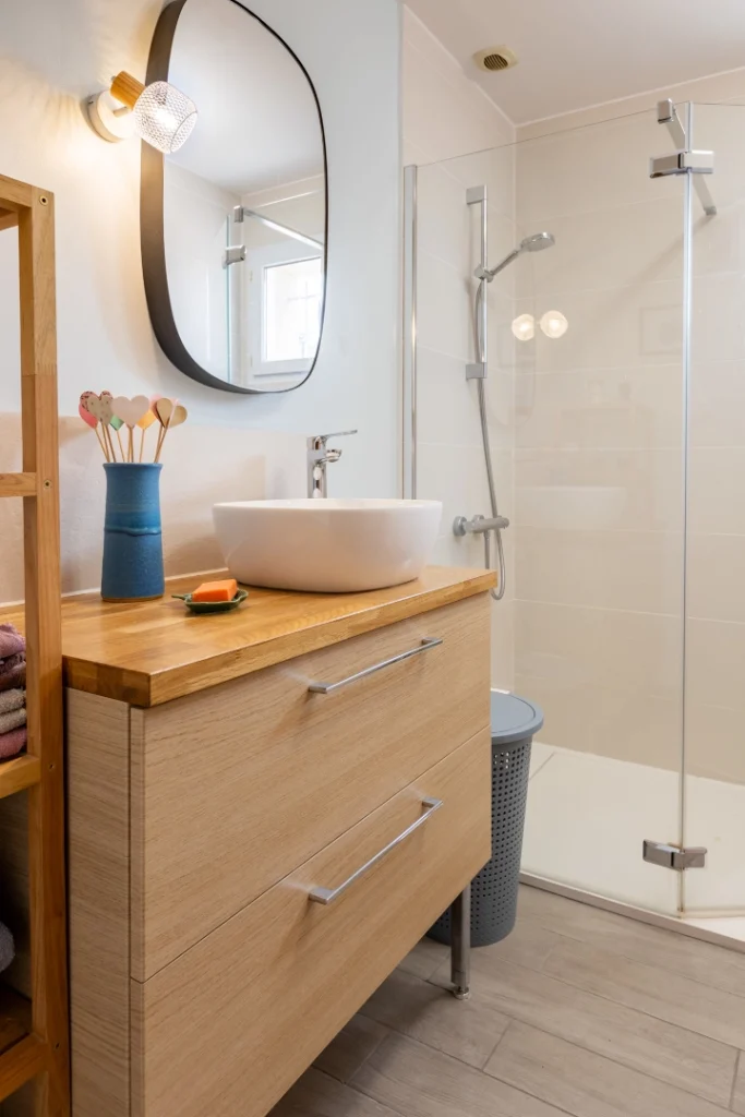 Salle de bain moderne lumineuse avec un meuble vasque en bois clair à deux grands tiroirs, surmonté d'une vasque ronde blanche. À côté, une douche à l'italienne avec paroi vitrée transparente et un sol en carrelage imitation parquet gris.