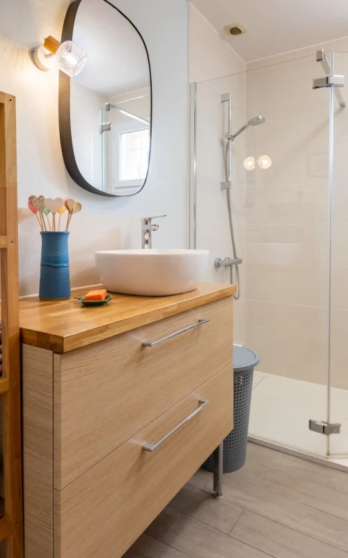 Salle de bain moderne lumineuse avec un meuble vasque en bois clair à deux grands tiroirs, surmonté d'une vasque ronde blanche. À côté, une douche à l'italienne avec paroi vitrée transparente et un sol en carrelage imitation parquet gris.
